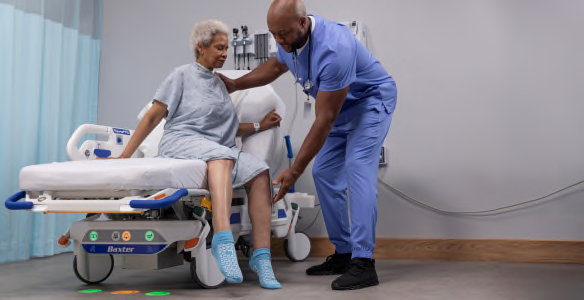 A caregiver assists a patient seated at the edge of a Dynamo Series stretcher